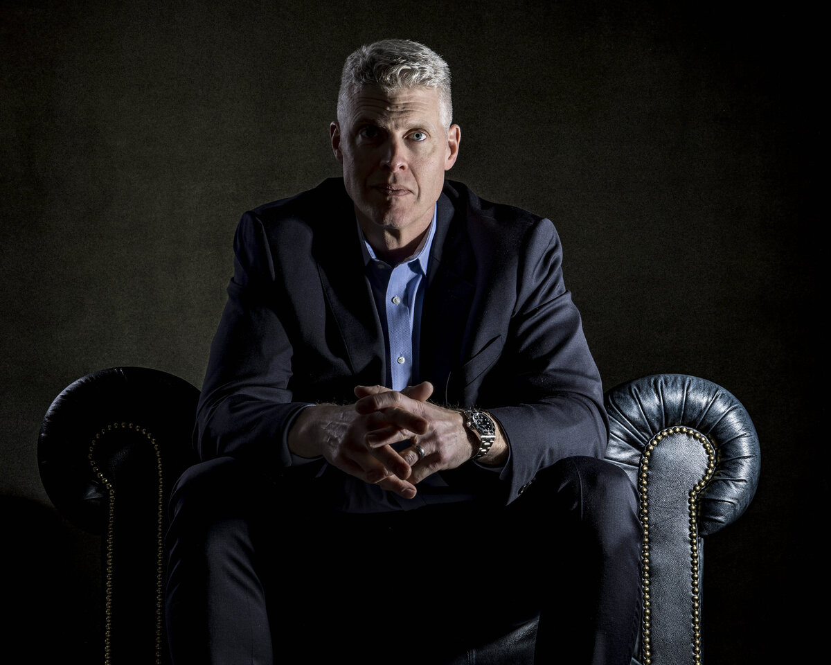 Professional headshot in CT of man in suit seated on gray leather chair, posed with hands together by Rob Faber Photography.