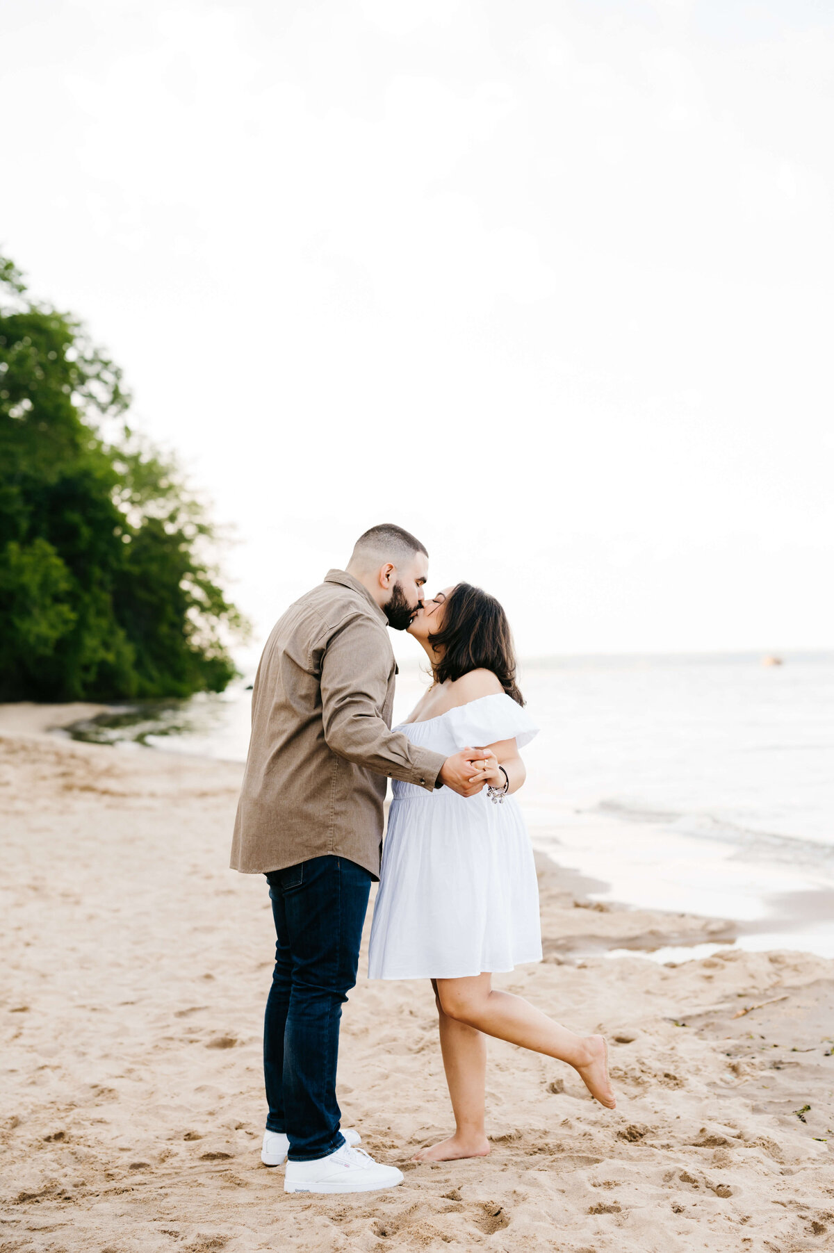 Virginia wedding photographer captures man and woman kissing as they hold hands and the woman pops up her foot on the beach during engagement photos