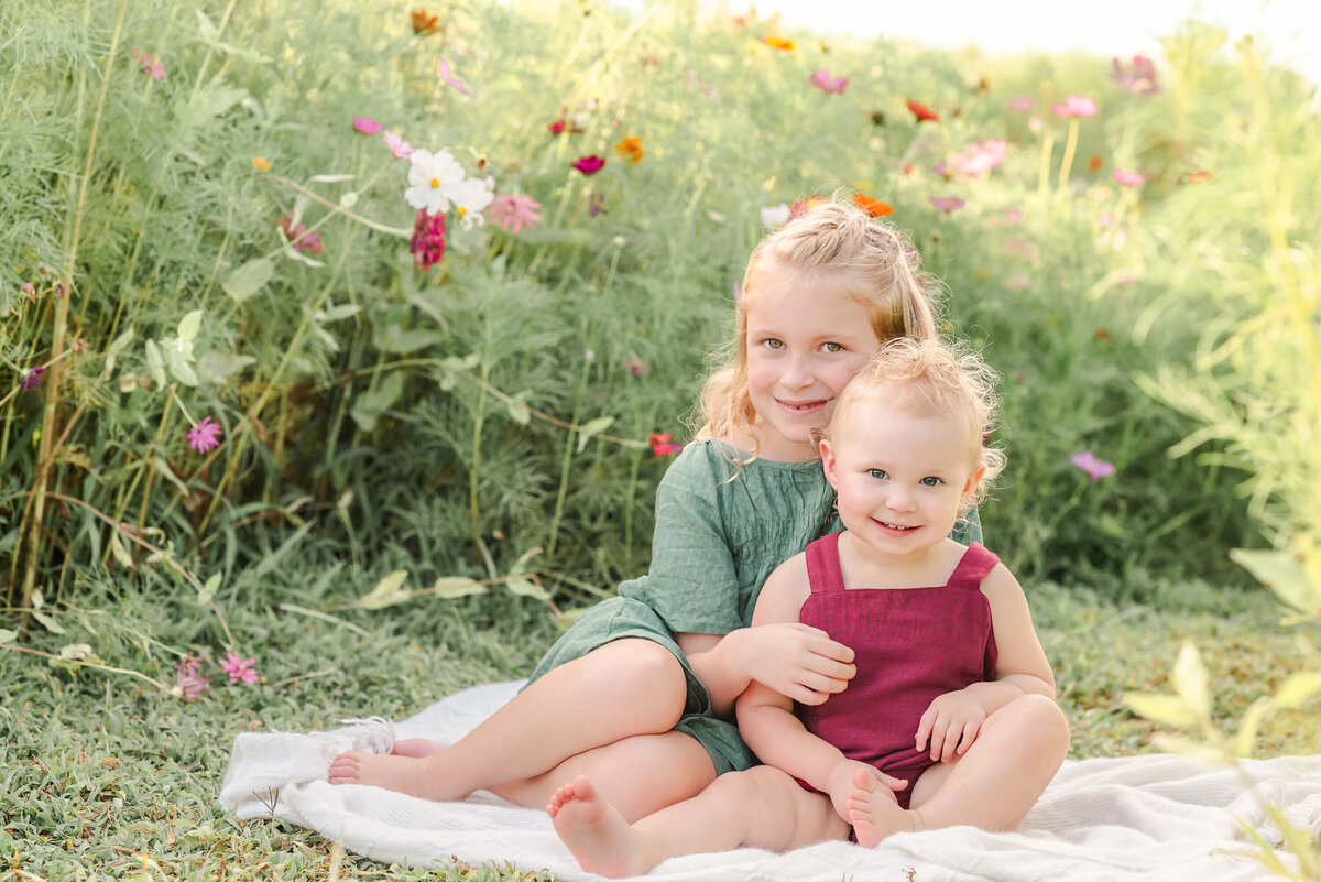 A pair of blond sisters sits on a white blanket in a flower field.