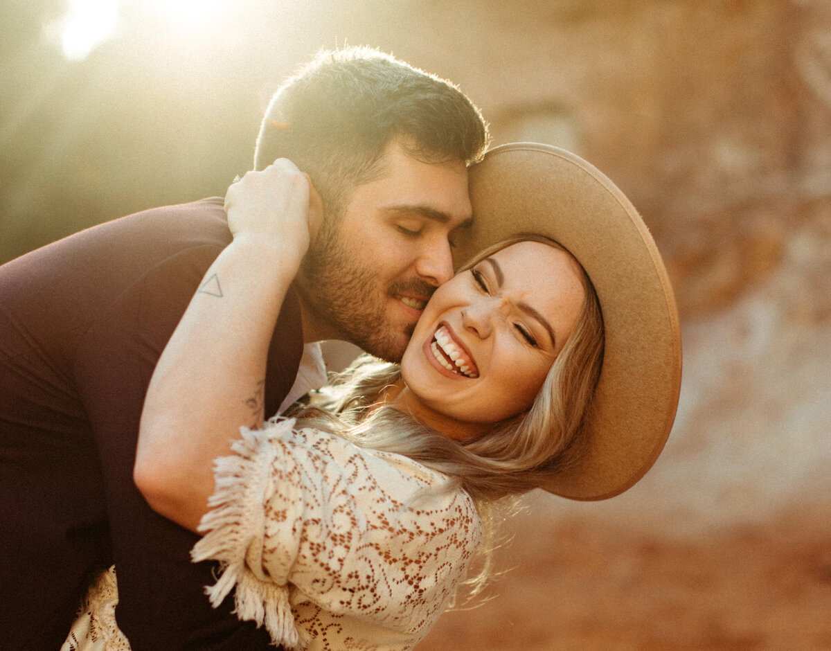 Groom making his bride laugh as he leans her back and kisses her cheek with golden hour sunset lighting behind them