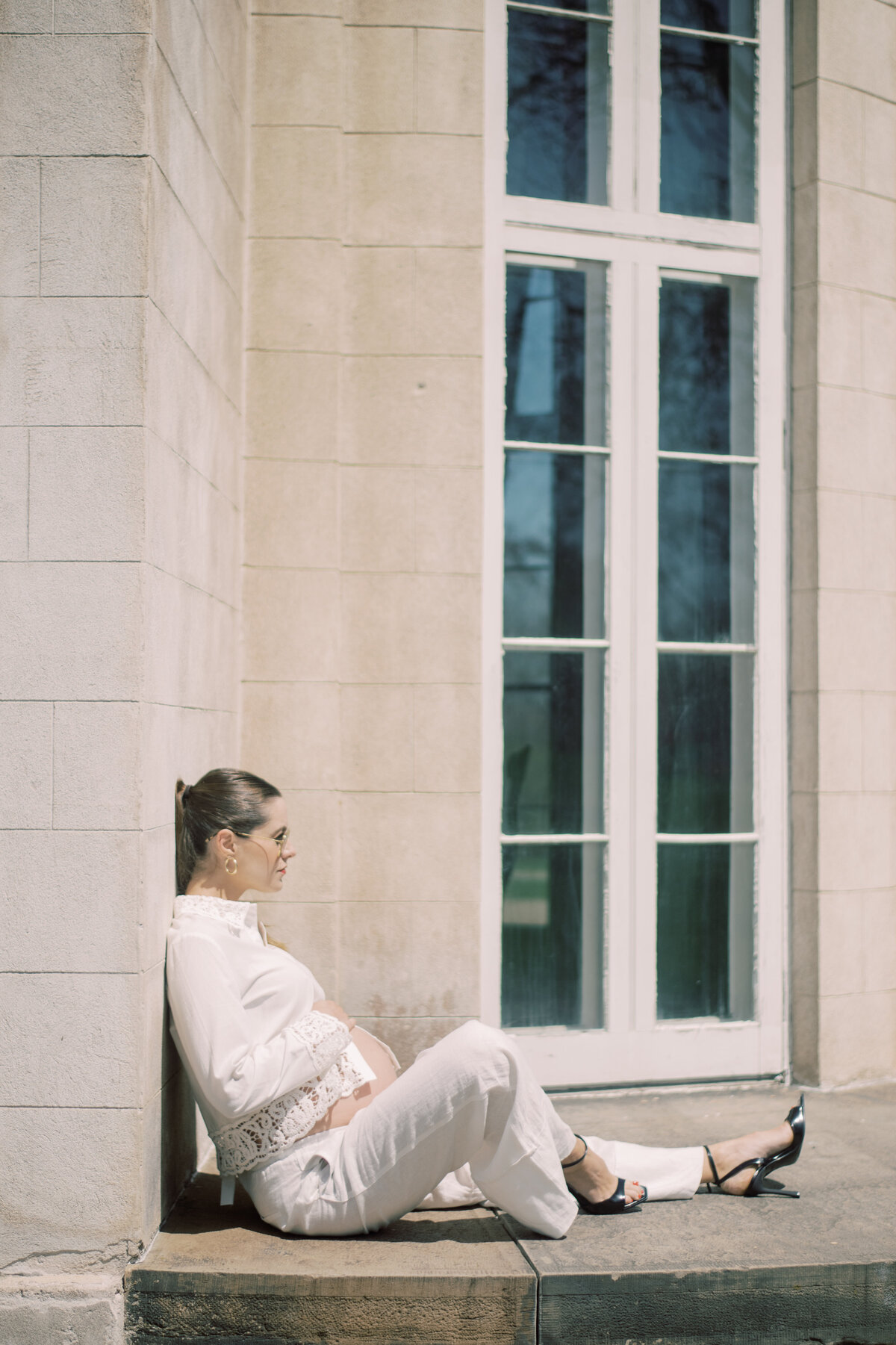 Graceful editorial shot of a maternity subject in heels and white linen, hand extended to the column in subtle motion