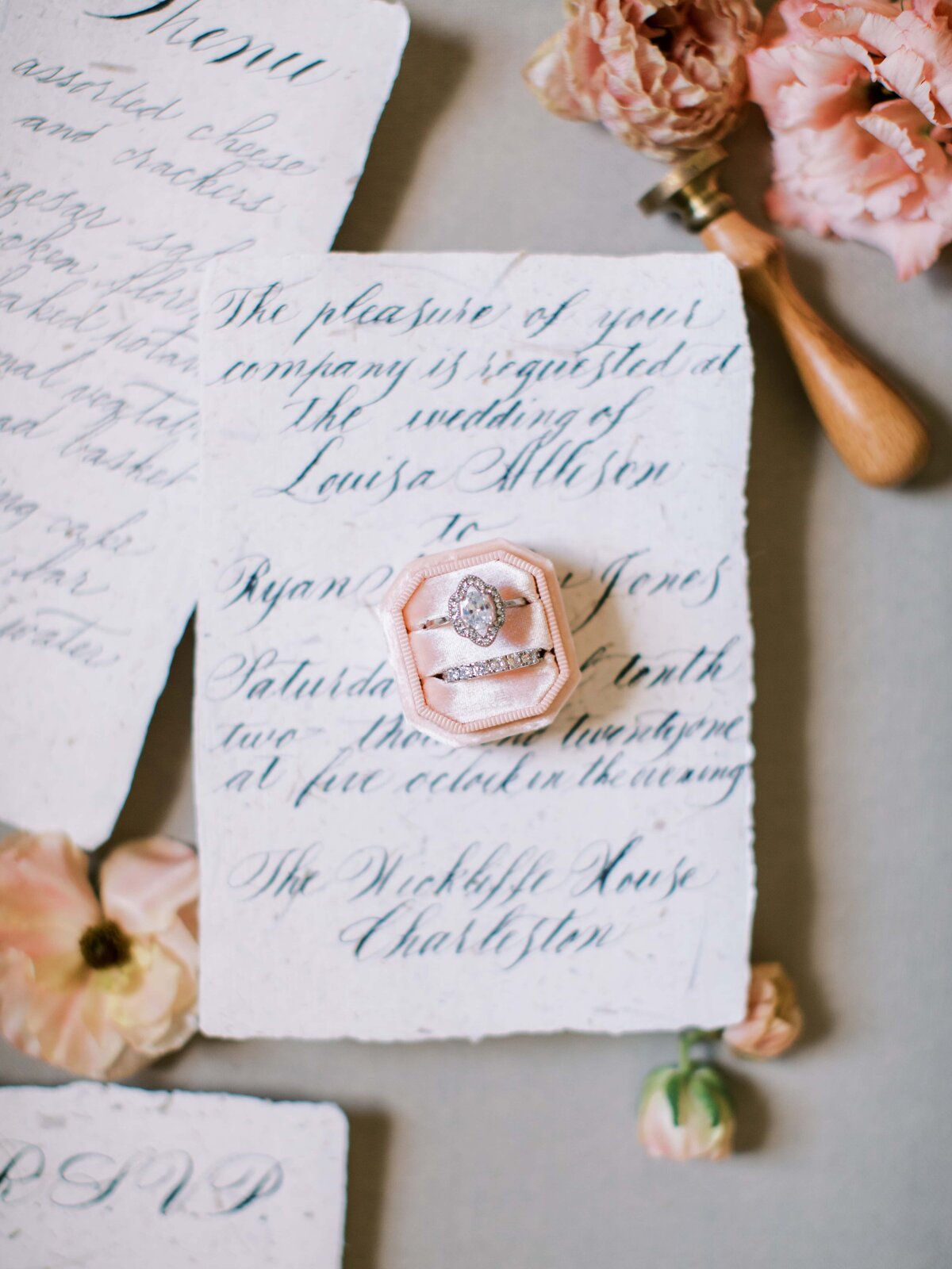 Image of a wedding ring and engagement ring in a velvet pink ring box on a hand written wedding invitation with pink blooms around it