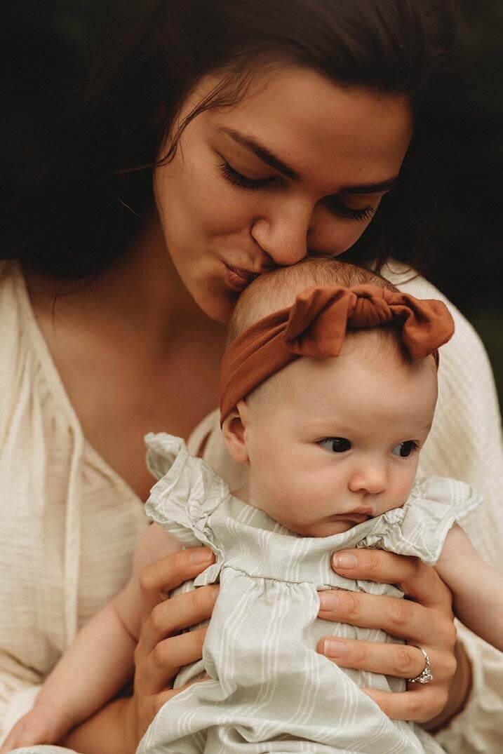a mom holds her baby on her lap while she kisses the top of her head in canandiagua ny