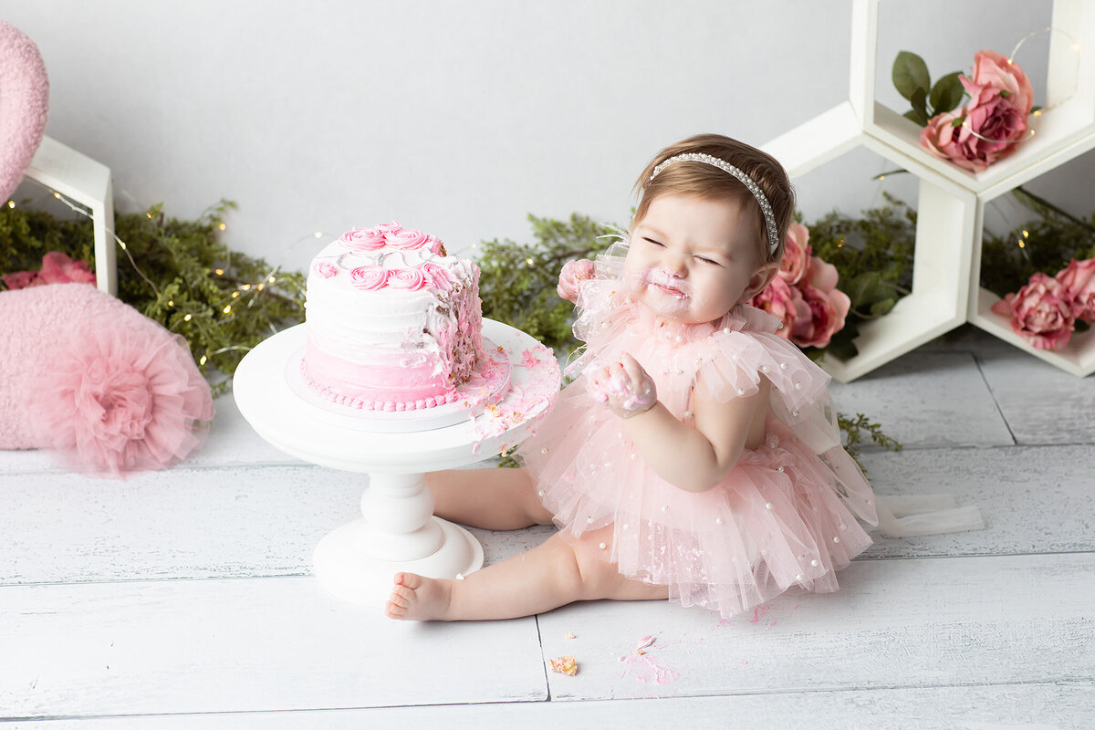 Dallas baby milestone photographer showcases a joyful cake smash moment as a baby girl in a pearl-accented pink dress scrunches her face with delight, frosting on her cheeks and hands beside a pink ombré cake.