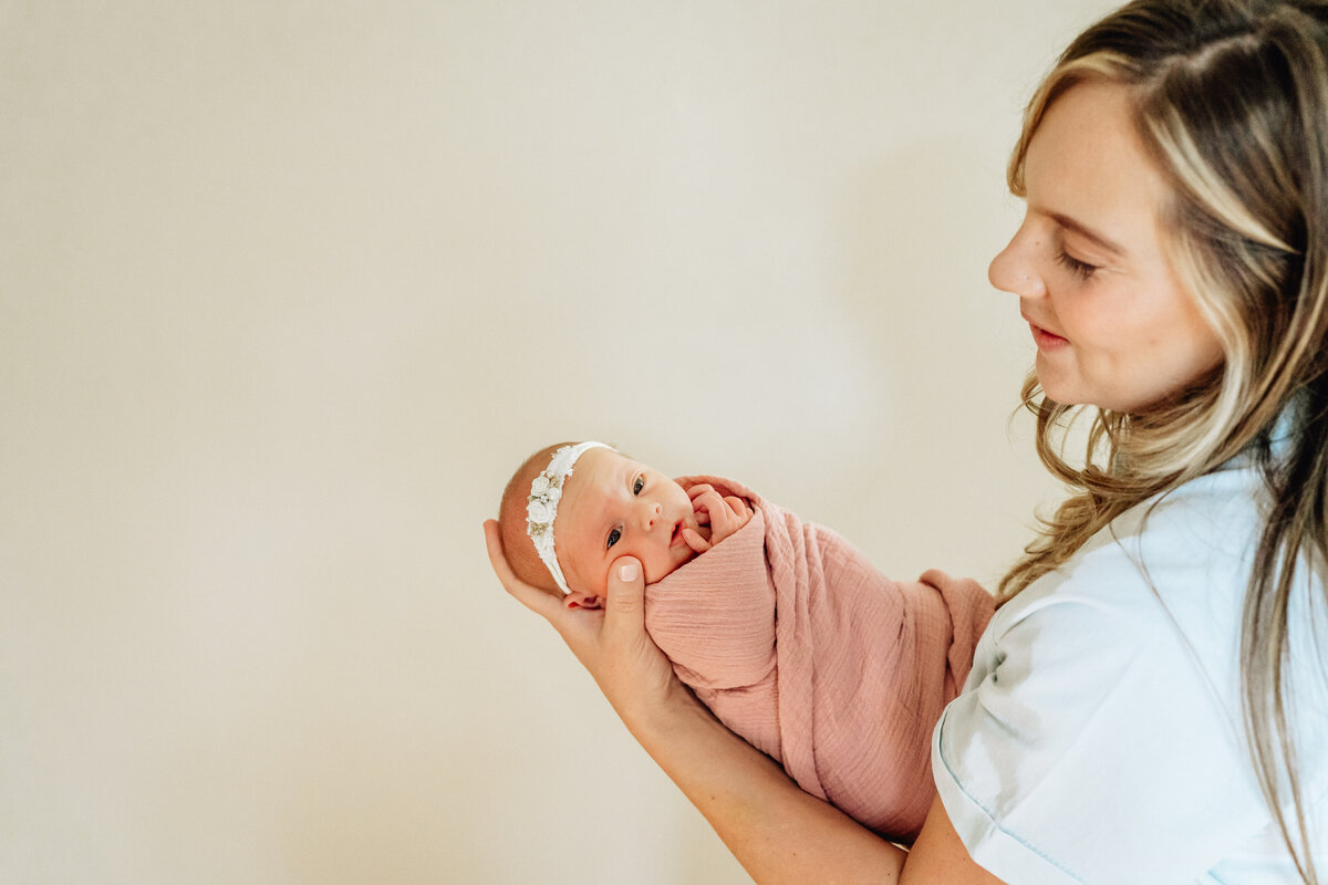 A mother holding her newborn baby wrapped in a pink swaddle, against a simple, light-colored background.