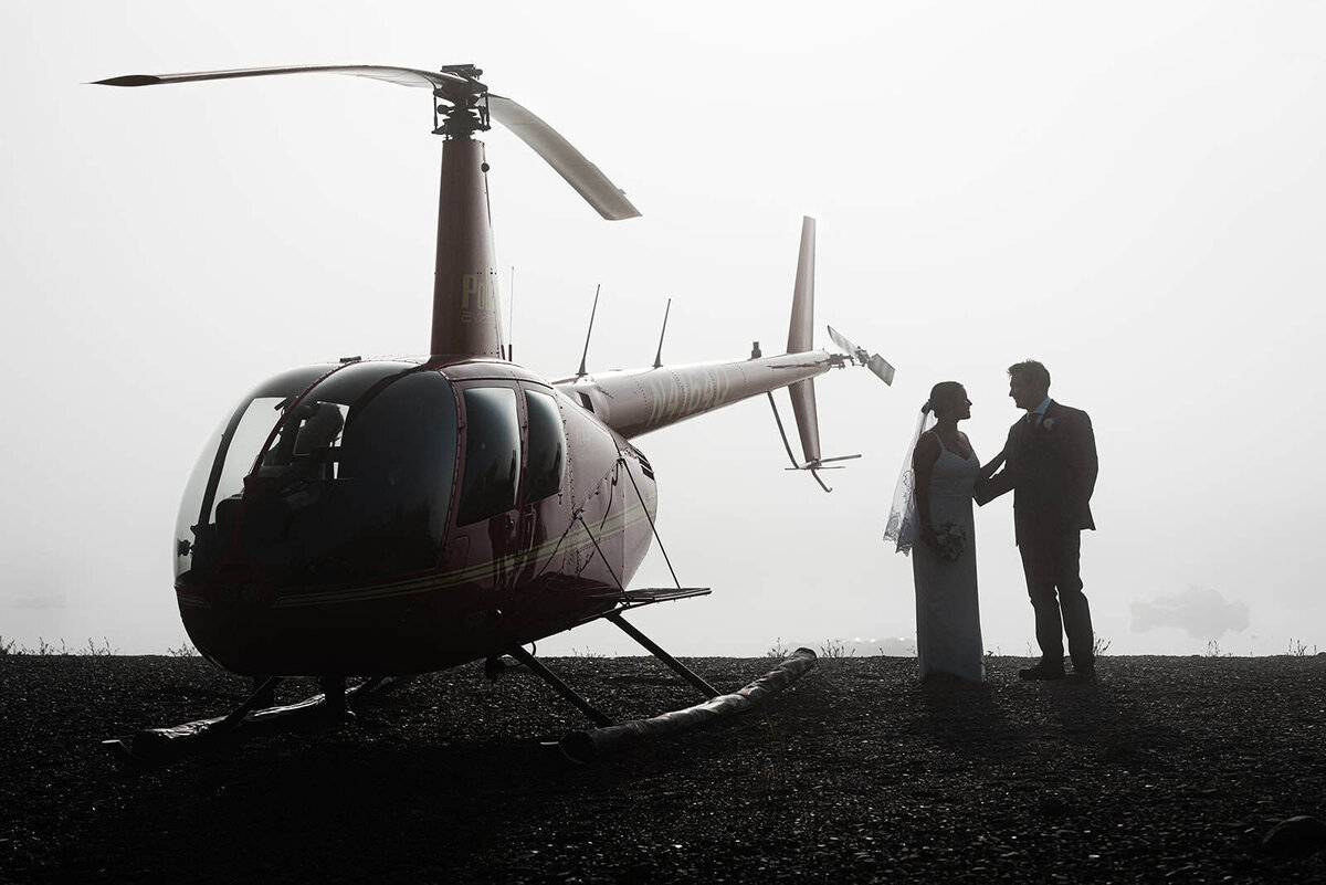 A bride and groom disembark from a helicopter, and they and the side profile of the hlicopter are silhouetted in the fog coming off the glacier lagoon. they are coming from exchanging vows during their Alaska private helicopter Elopement wedding in Seward Alaska, and are gently embracing as they look at each other.  