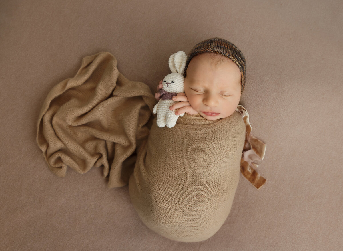 Baby boy in a taupe swaddle is holding a bunny for a brooklyn ny newborn photoshoot. Captured by top Brooklyn newborn photographer Chaya Bornstein