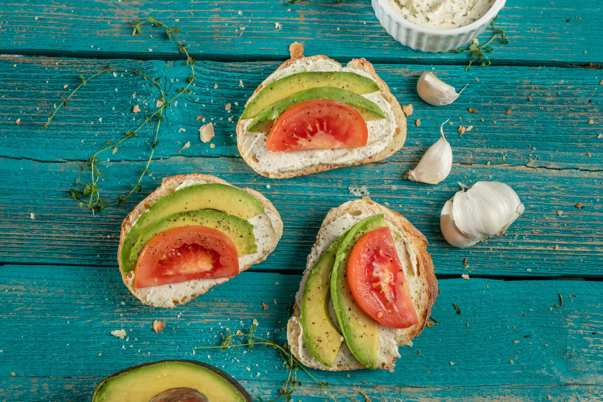sourdough avocado toast with alouette garlic and herb cheese spread and tomato wedges on a blue rustic table.