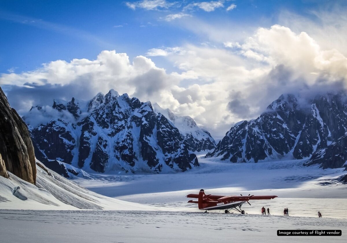 Images of a red and white ski plane landed on the snowfield glacier in Denali National Park.  There are people standing under the right wing of the plane--they look tiny next to the plane, with huge, snow covered craggy mountains shooting up behind and around them out of the snowy glacier.  The mountains fill nearly the entire back of the frame, except for a tiny peek of bluebird blue sky between the Alaska mountain peaks, a scene perfect for a non-helicopter glacier elopement in Alaska. 