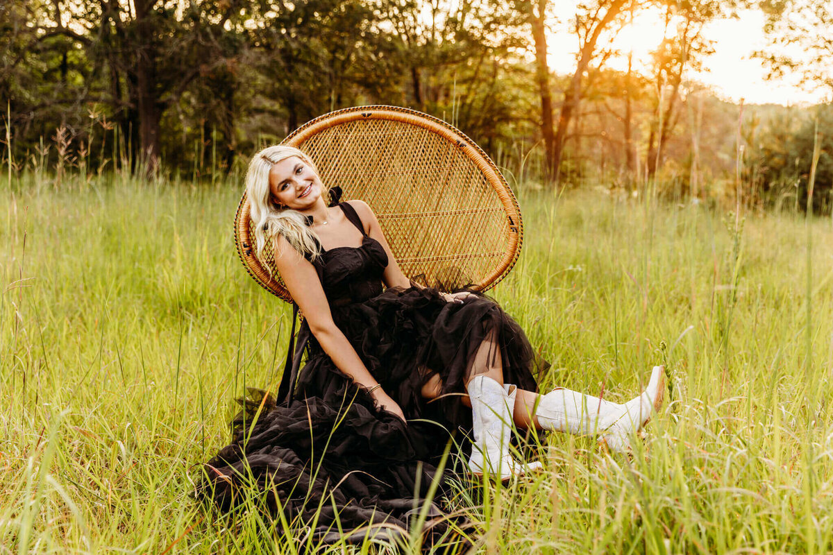 stunning teen girl in a black gown sitting on a peacock chair in a field at sunset as she kicks out her white cowboy boots and smiles captured by Ashley Kalbus.