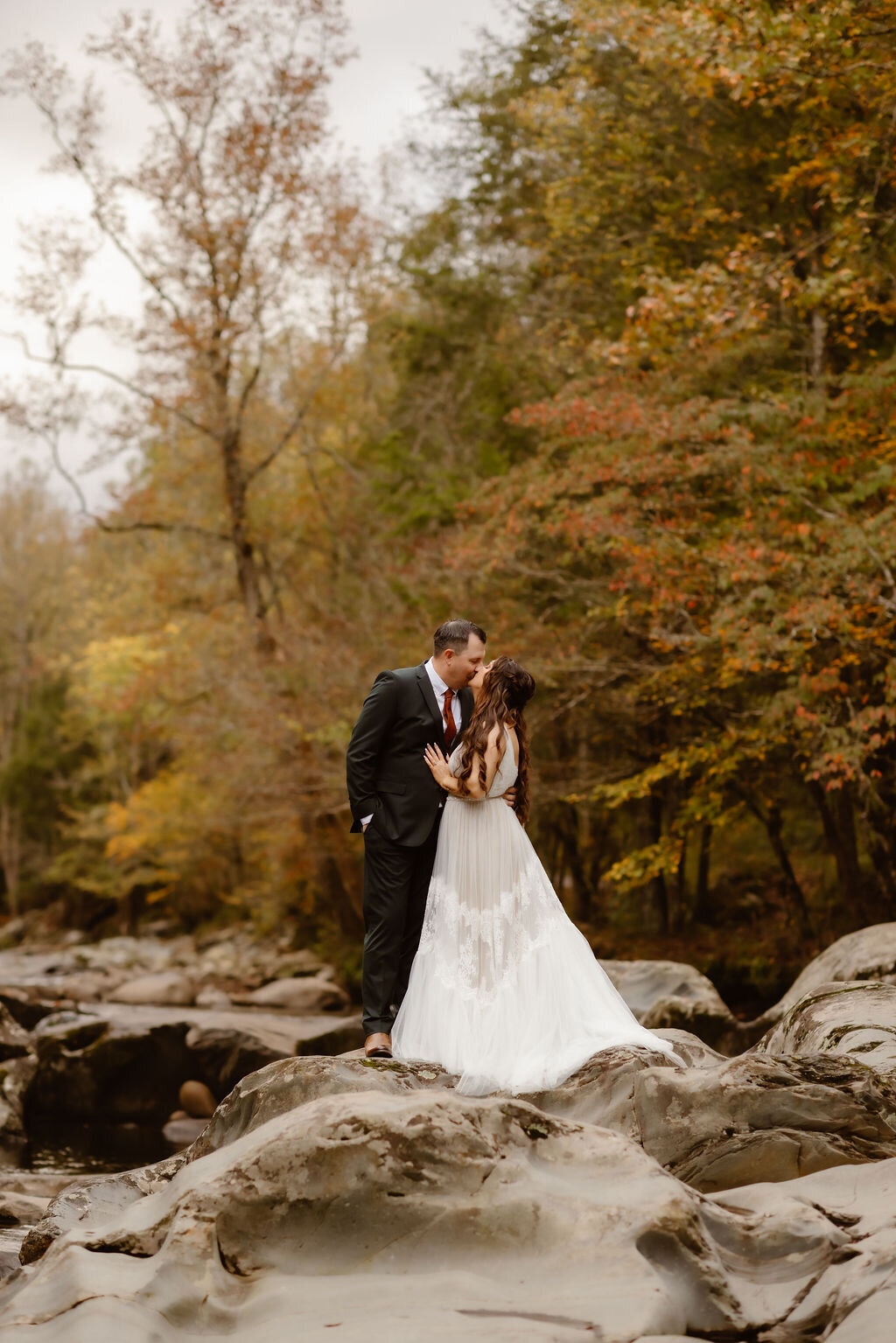 Bride and groom sharing a romantic kiss atop the smooth river rocks at Greenbrier, framed by golden autumn leaves during their eloping to Gatlinburg celebration.
