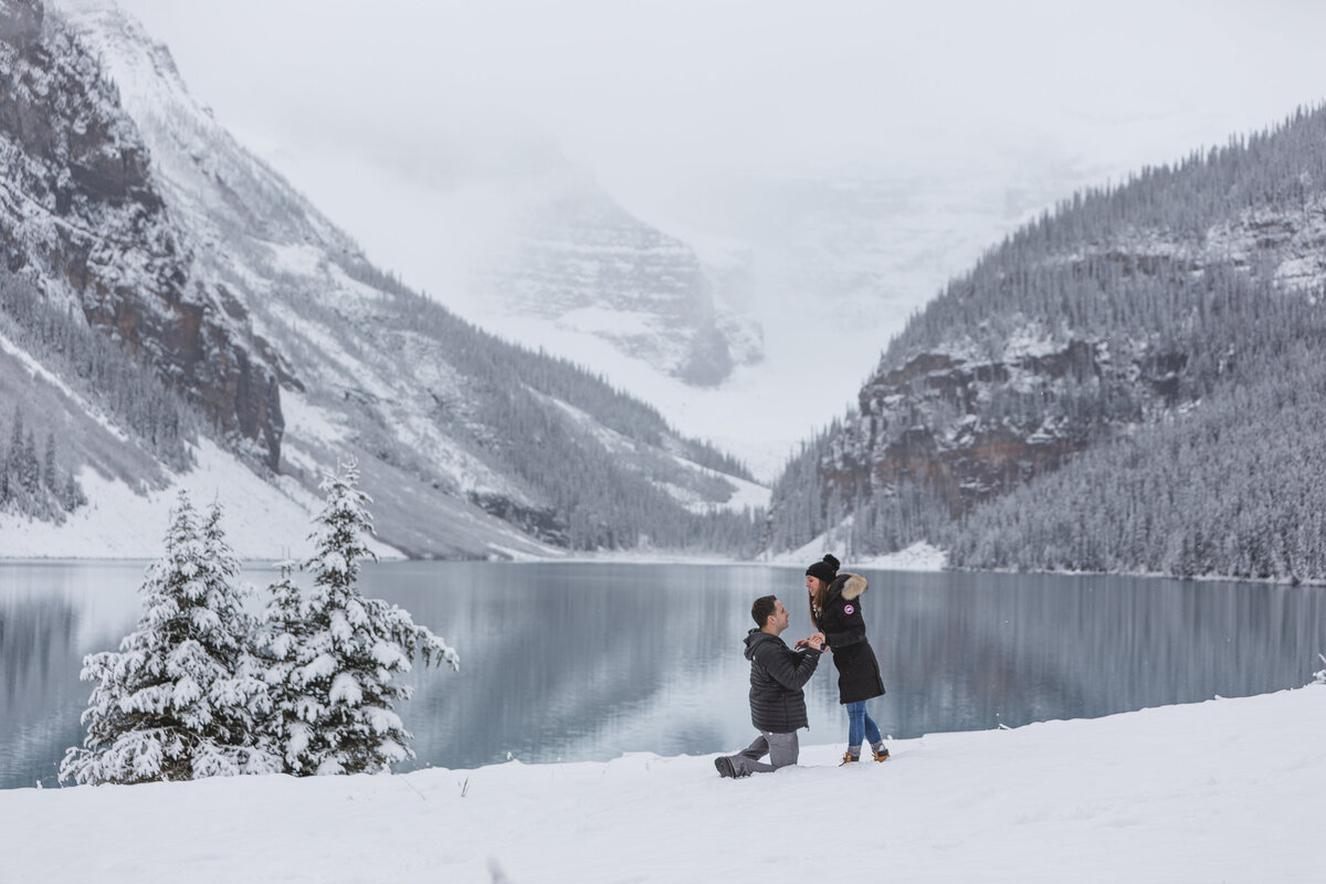 Couple embrases by the frozen lake in Lake Louise with misty mountain views, perfect for a magical Banff proposal photo session.