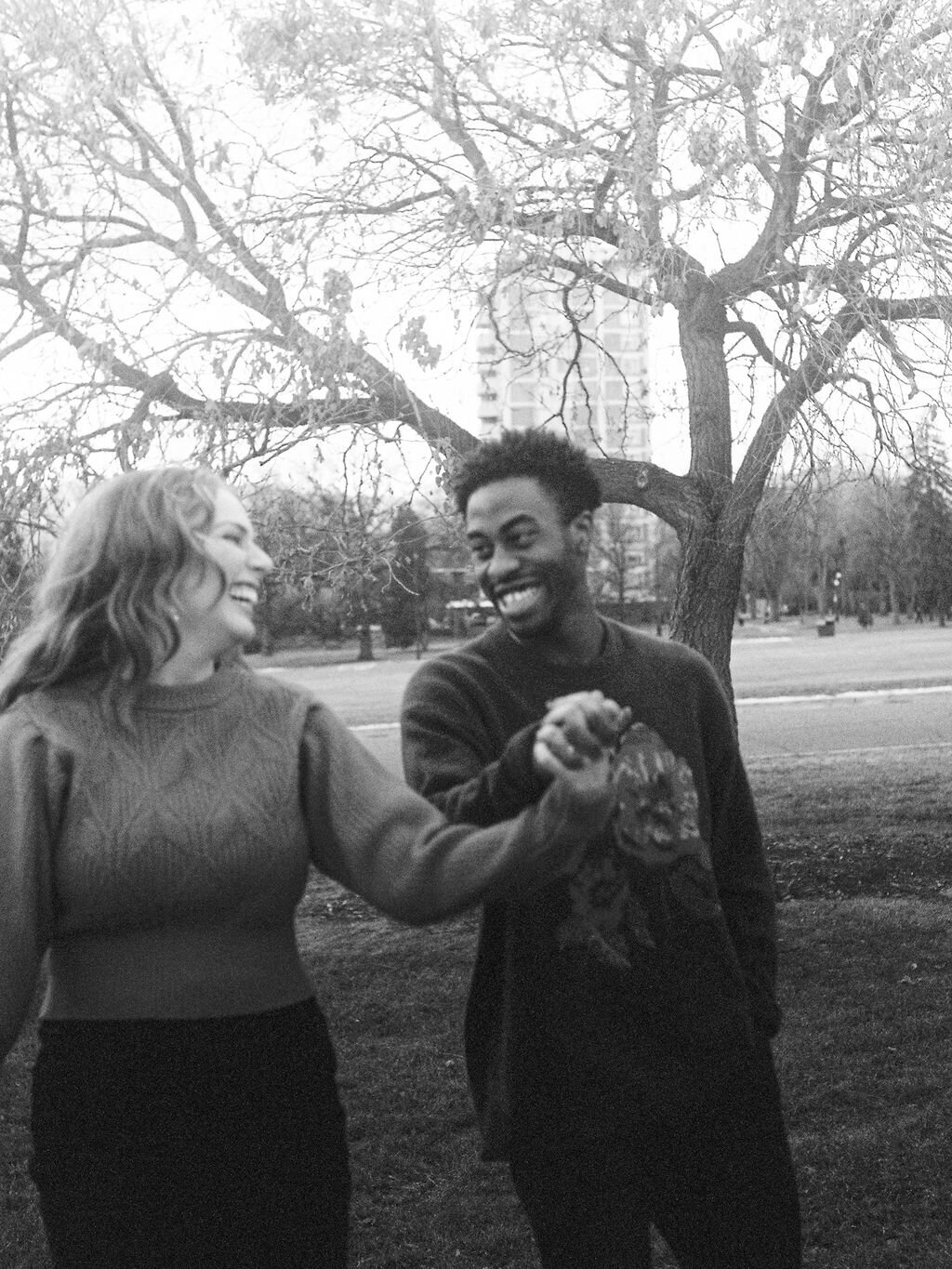 A couple holding hands walks through Cheesman Park. They are looking at each other.