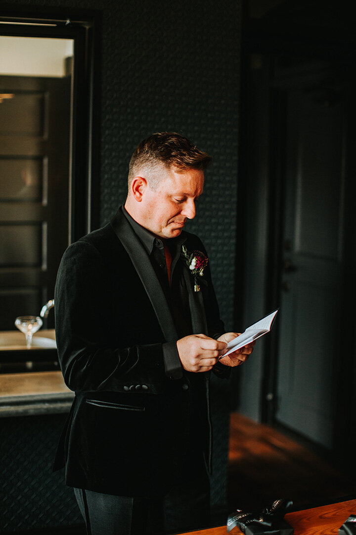 Groom wearing a black tuxedo holding a card stock wedding booklet contemplates while he reads in a room.