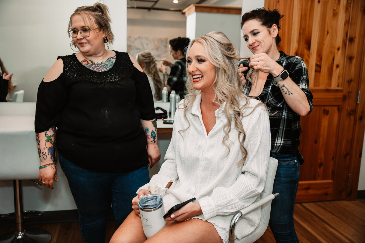 A smiling bride sits in a chair while her hair is worked on in white pajamas