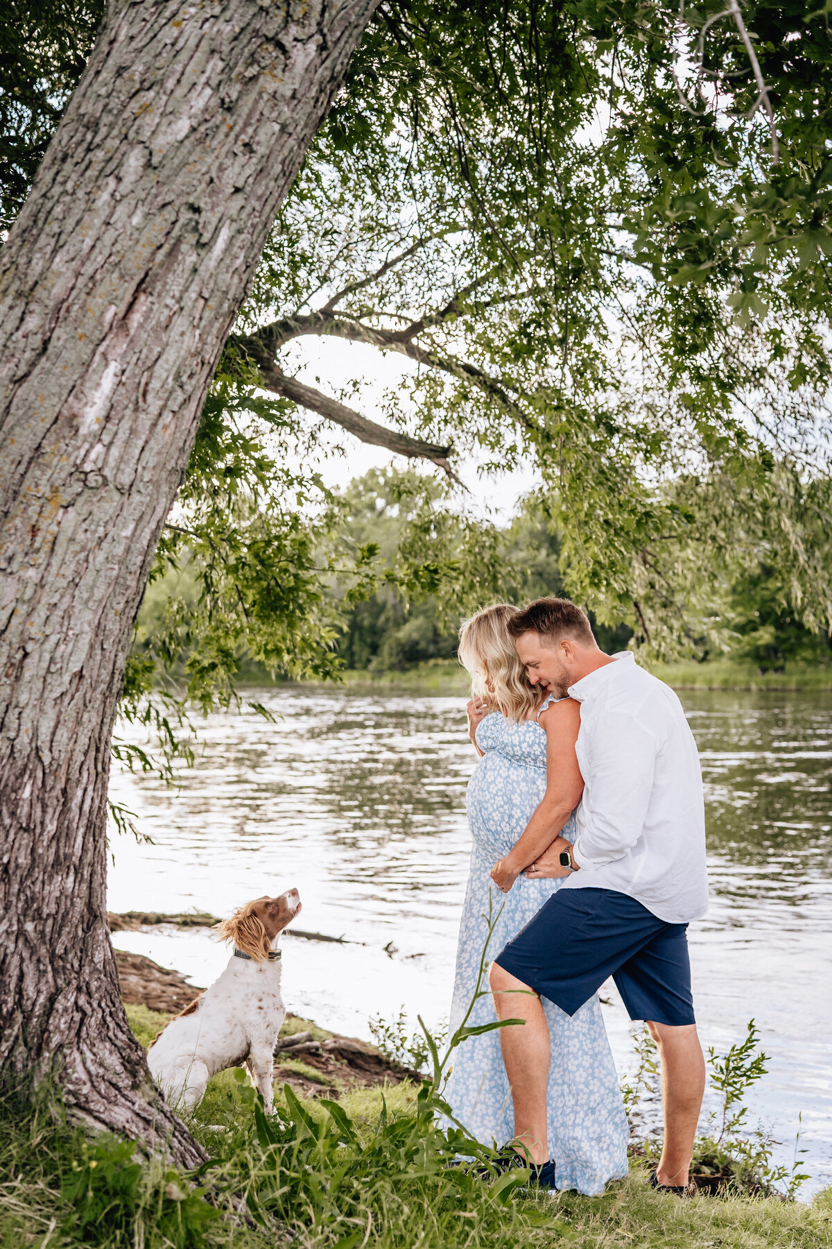 A couple stands near a river, embracing each other while a dog sits nearby, looking up at them. The woman, wearing a light blue floral dress, is visibly pregnant, and the man is wearing a white shirt and navy shorts.