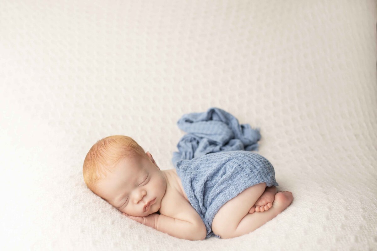 Tiny newborn boy with soft red hair posed with his hands under his chin on a dreamy fabric backdrop