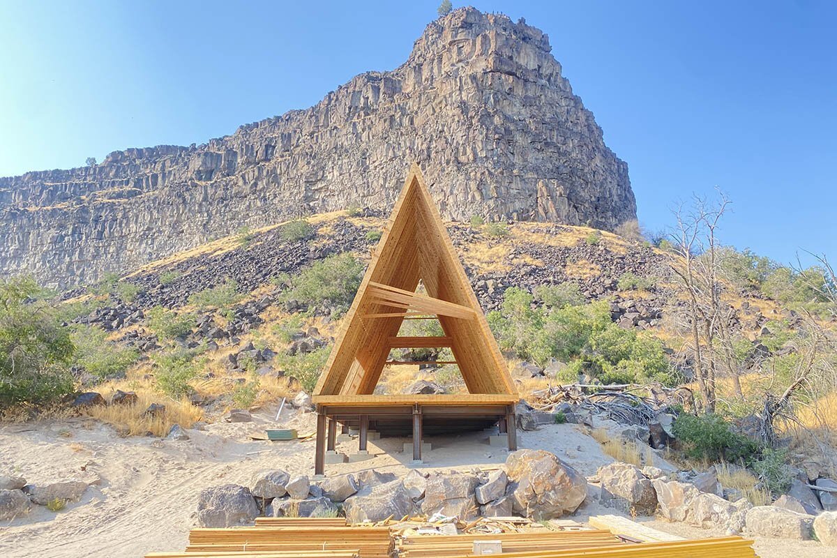 An A-frame cabin under construction at The Perrine Hideaway, showing exposed wooden beams and an open-frame structure against a scenic canyon