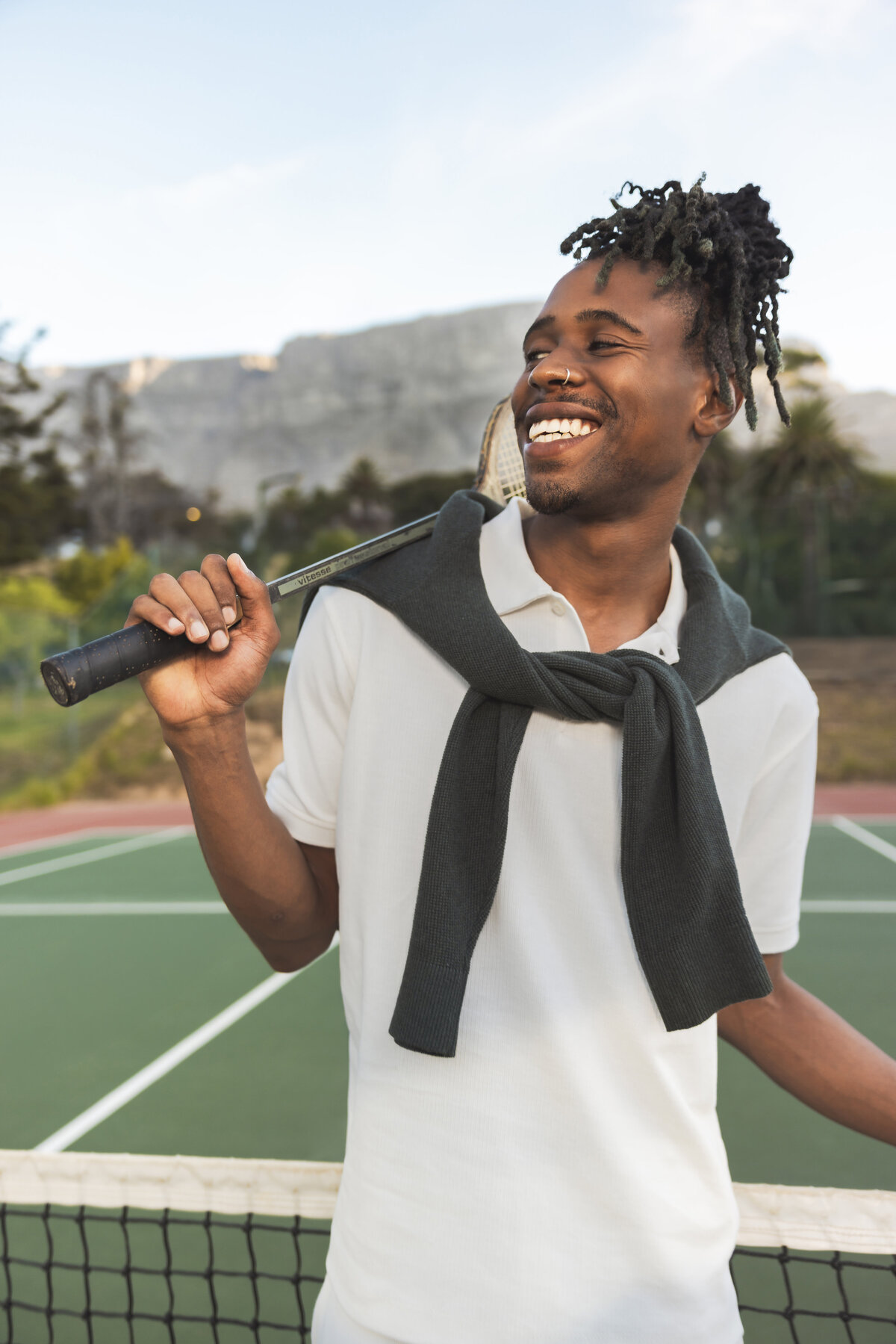 Smiling man in a white tennis outfit with a dark sweater draped over his shoulders, holding a racket behind his neck, standing on a tennis court with Table Mountain in the background.