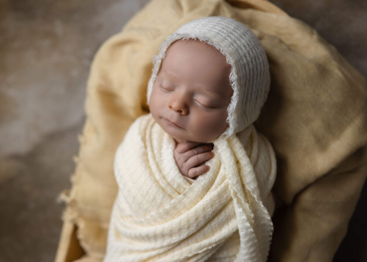 close up of newborn baby in cream wrap and bonnet asleep in wooden bowl