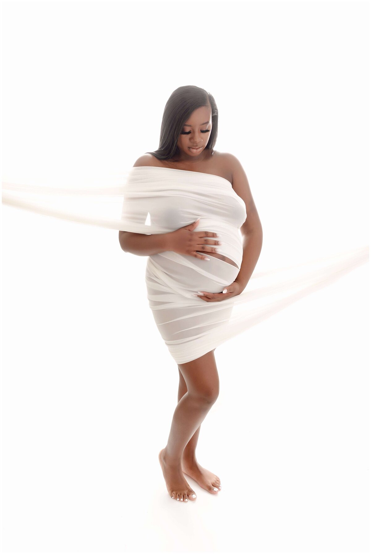 a pregnant person in a white fabric holding her belly and looking down admiring..