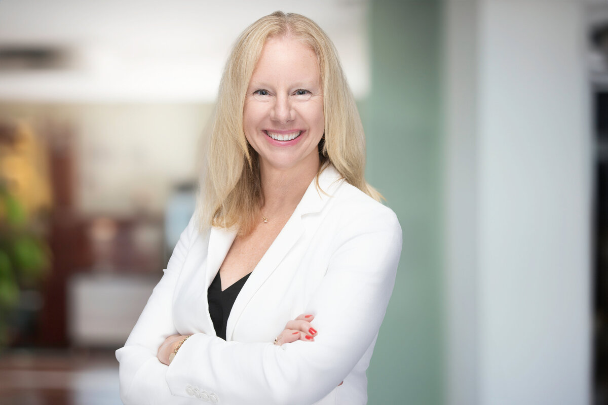 Professional headshot of a smiling blonde woman in a white blazer with arms crossed, captured at Janel Lee Photography studio in downtown Cincinnati, Ohio. She stands against a softly blurred modern office backdrop, conveying confidence, warmth, and approachability—ideal for corporate headshots and personal branding sessions
