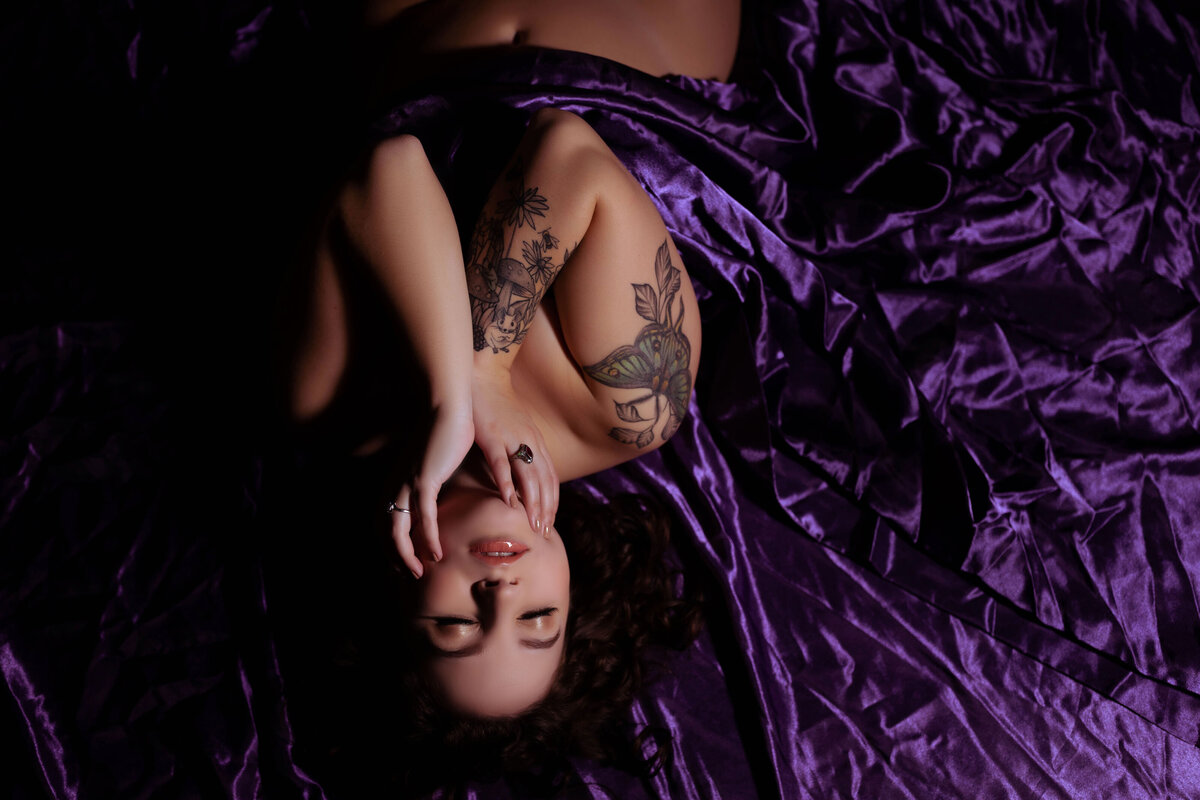 Boudoir Photographer, a woman lays on bed wrapped only in purple sheets, her tattoos exposed