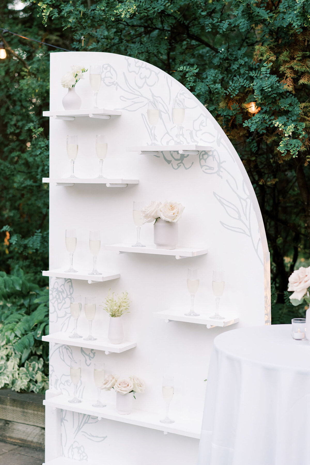 A white, semi-circular display stand with shelves holding champagne glasses and vases with flowers, set outdoors by greenery at a modern &amp; romantic wedding in Calgary. A round table with a white tablecloth is partially visible in the foreground, enhancing the elegance of this special day.