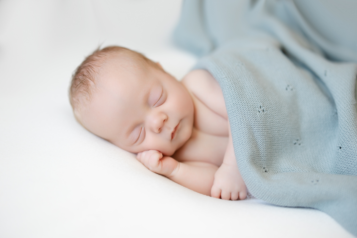 a newborn baby boy sleeps on a white sheet with one arm under his chin and is wrapped in a blue blanket 