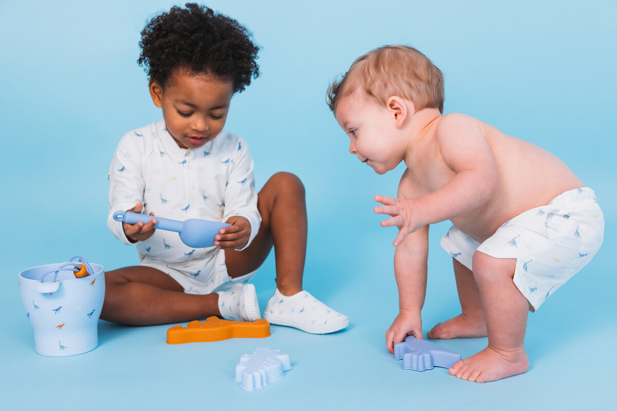 Two young children playing with beach toys, wearing matching Swim Essentials swimwear with dinosaur print, against a light blue background.