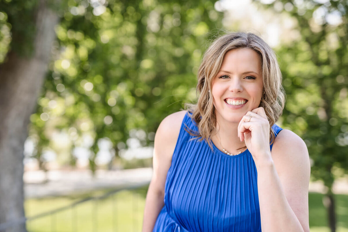 Happy women’s health doctor in pleated blue dress, leaning forward outdoors in Kelowna park.