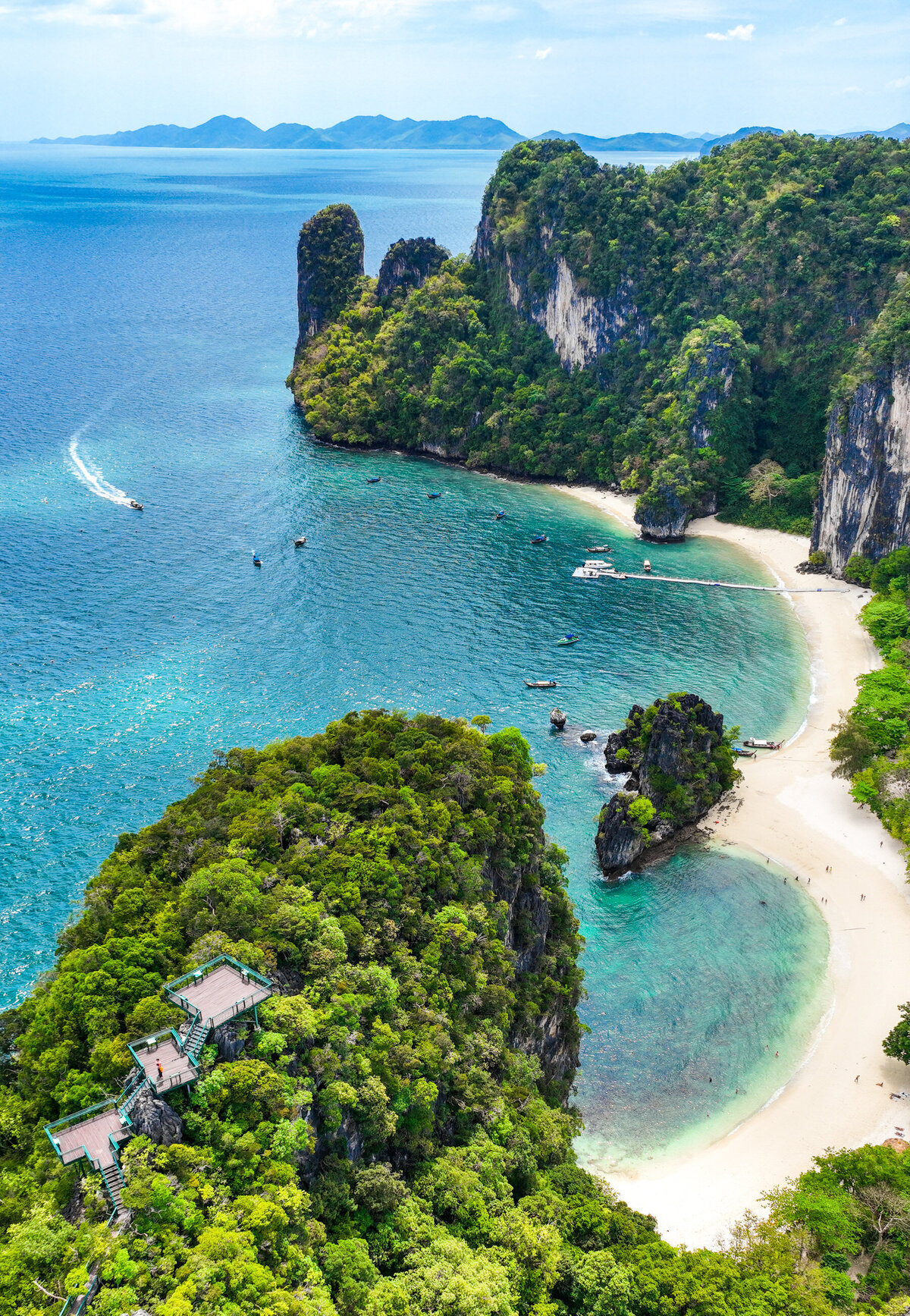 A stunning aerial view of a tropical island featuring lush greenery, sandy beaches, and turquoise waters. Boats are visible near the shore, and rocky cliffs rise dramatically alongside the sea. The horizon showcases distant islands under a clear blue sky.