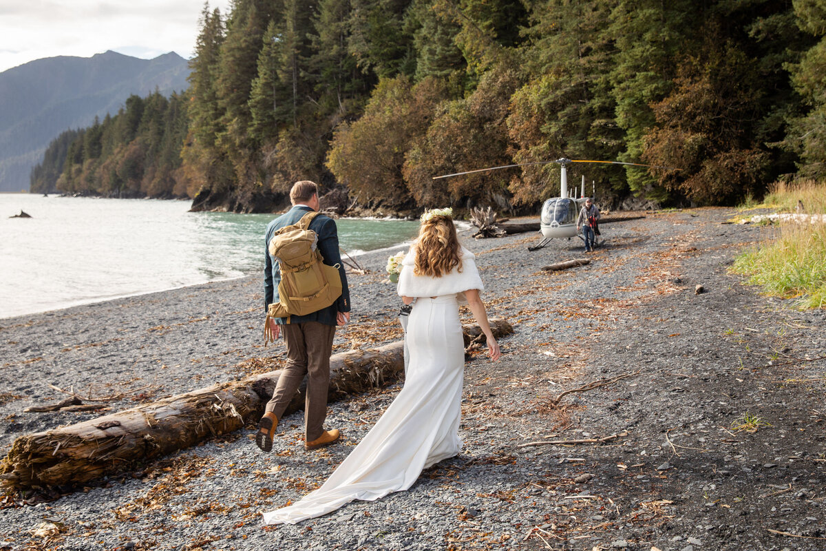A bride and groom walk along a beach in Alaska towards a helicopter waiting for them.