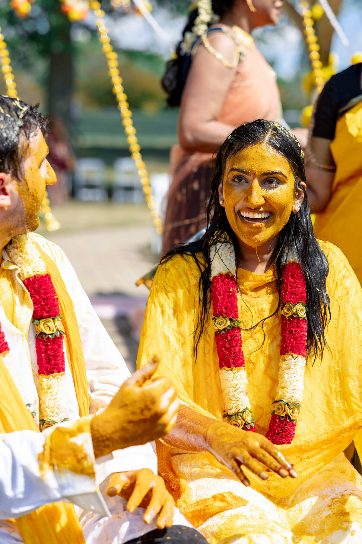 A couple participates in a traditional Indian pre-wedding ceremony, covered in turmeric paste. Both wear floral garlands of red and white roses. The scene is bright and festive with other participants blurred in the background.