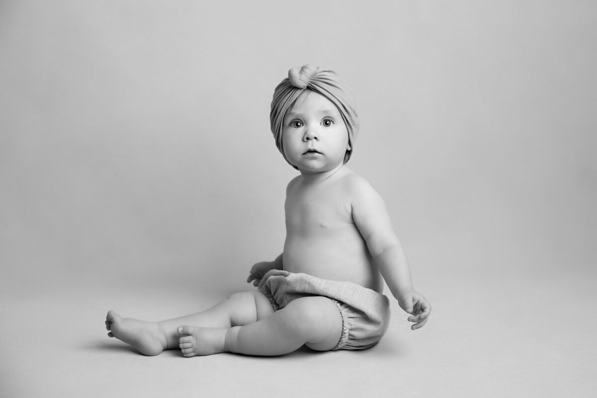 Side portrait of a baby girl sitting on a light background wearing bloomers and a knotted  head turban.