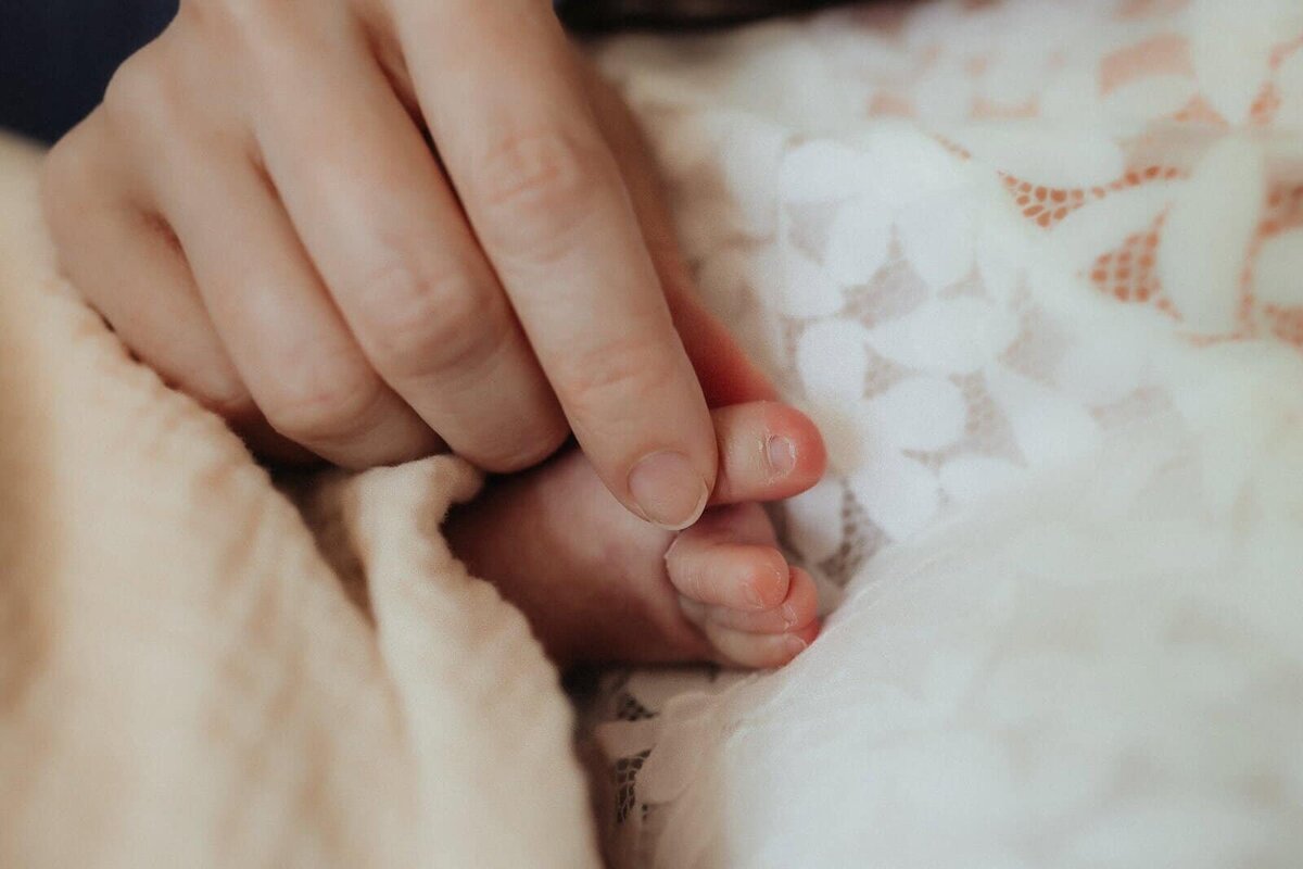 An adult hand gently holds a newborn baby's tiny foot, both resting on a soft, white lace blanket.