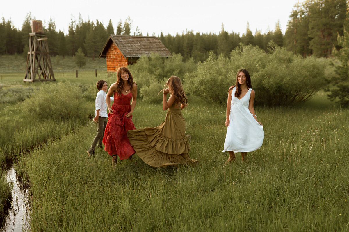 Mom and her three older kids are dancing in a beautiful grassy area with a small creek and rustic abandoned homestead behind them