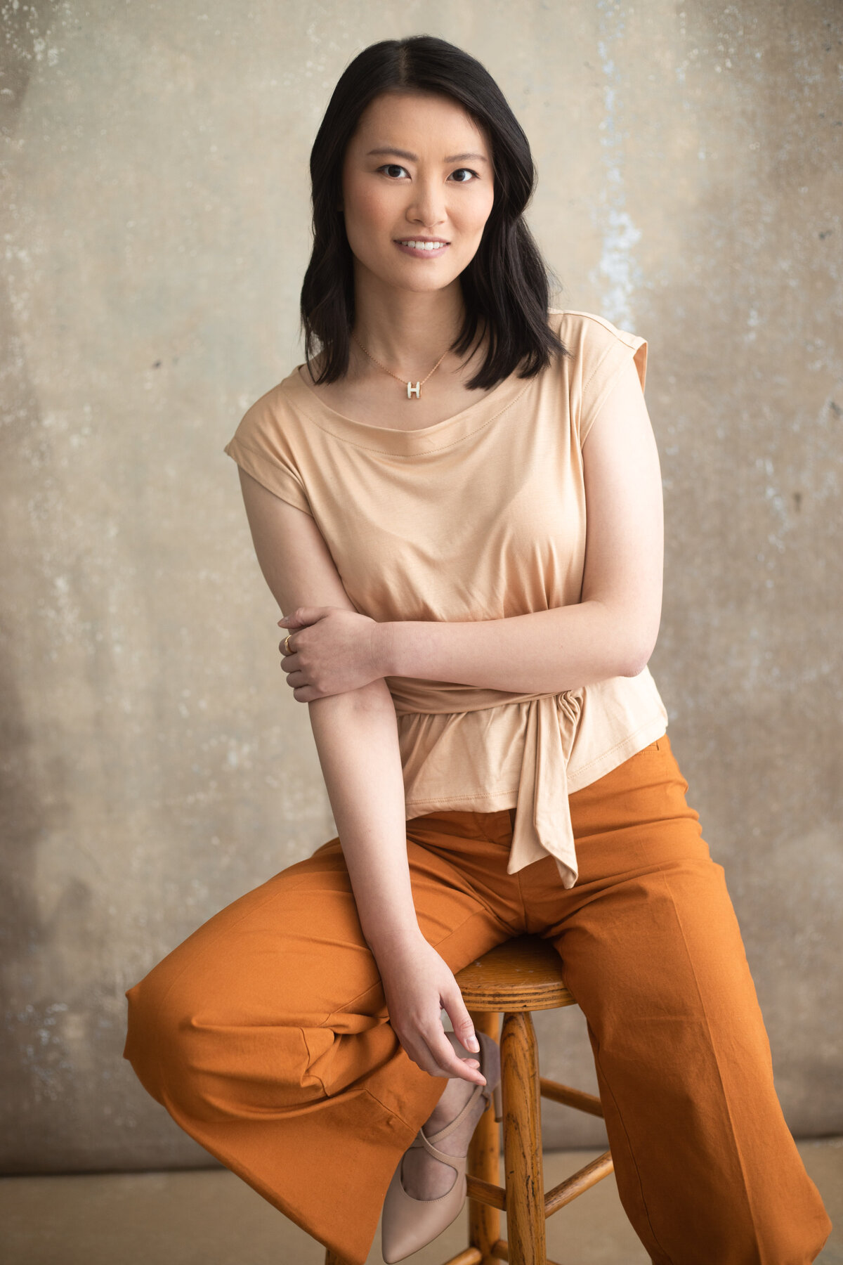 An Asian Chinese woman with orange pants and a light color shirt hold her arm and smiles for a portrait photo at Janel Lee Photography studios in Cincinnati Ohio
