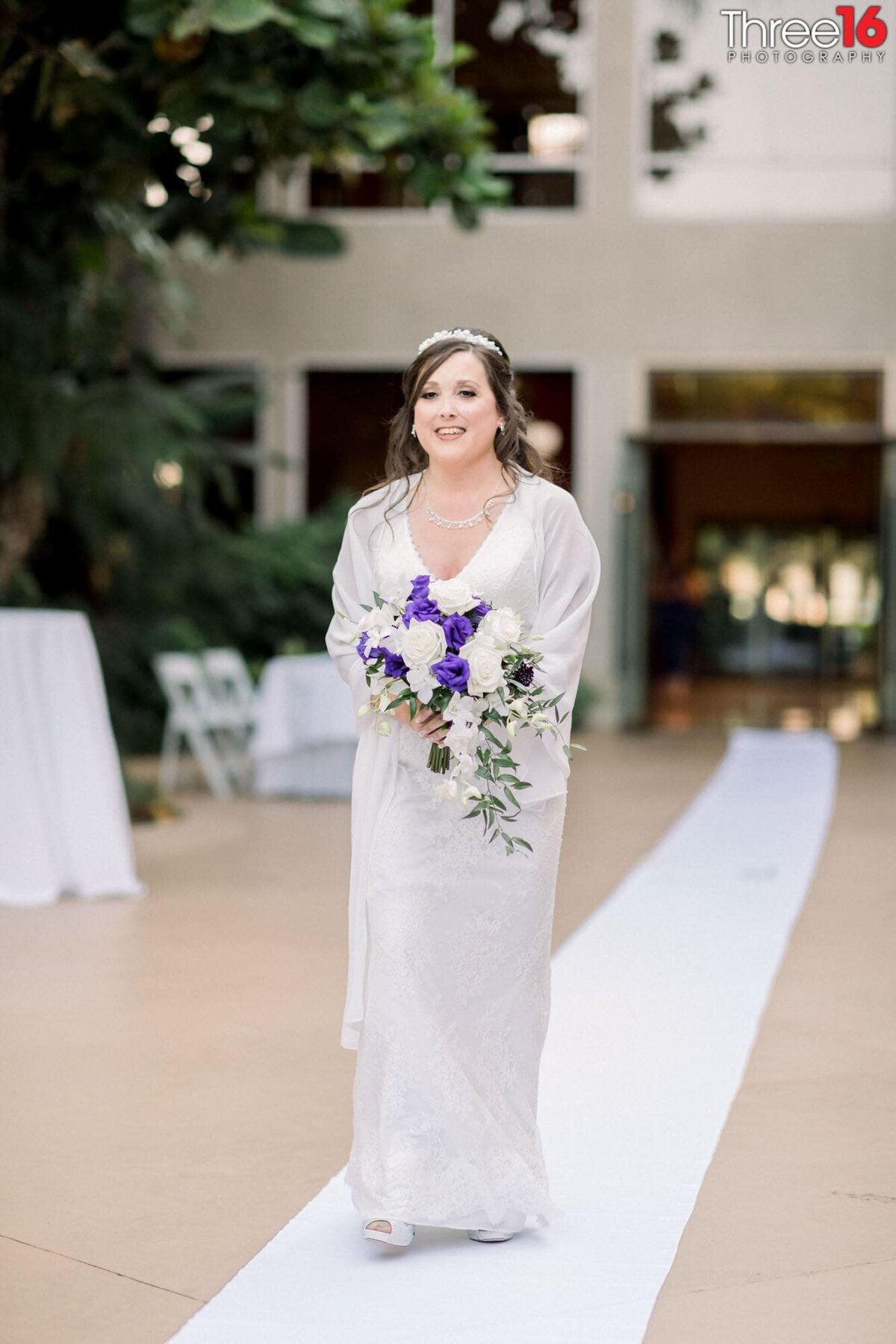 Smiling Bride walks up the aisle with purple & white bouquet in hand