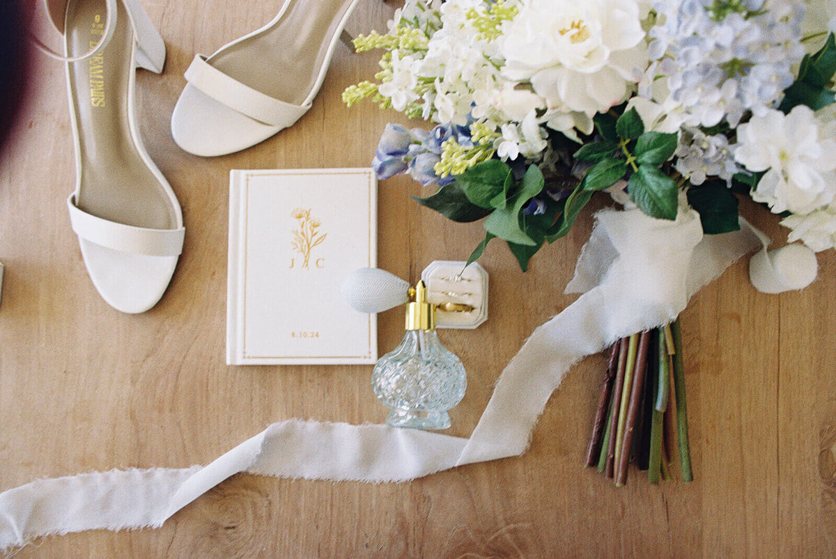Wedding flat lay featuring a bouquet, high heel shoes, invitation and perfume.