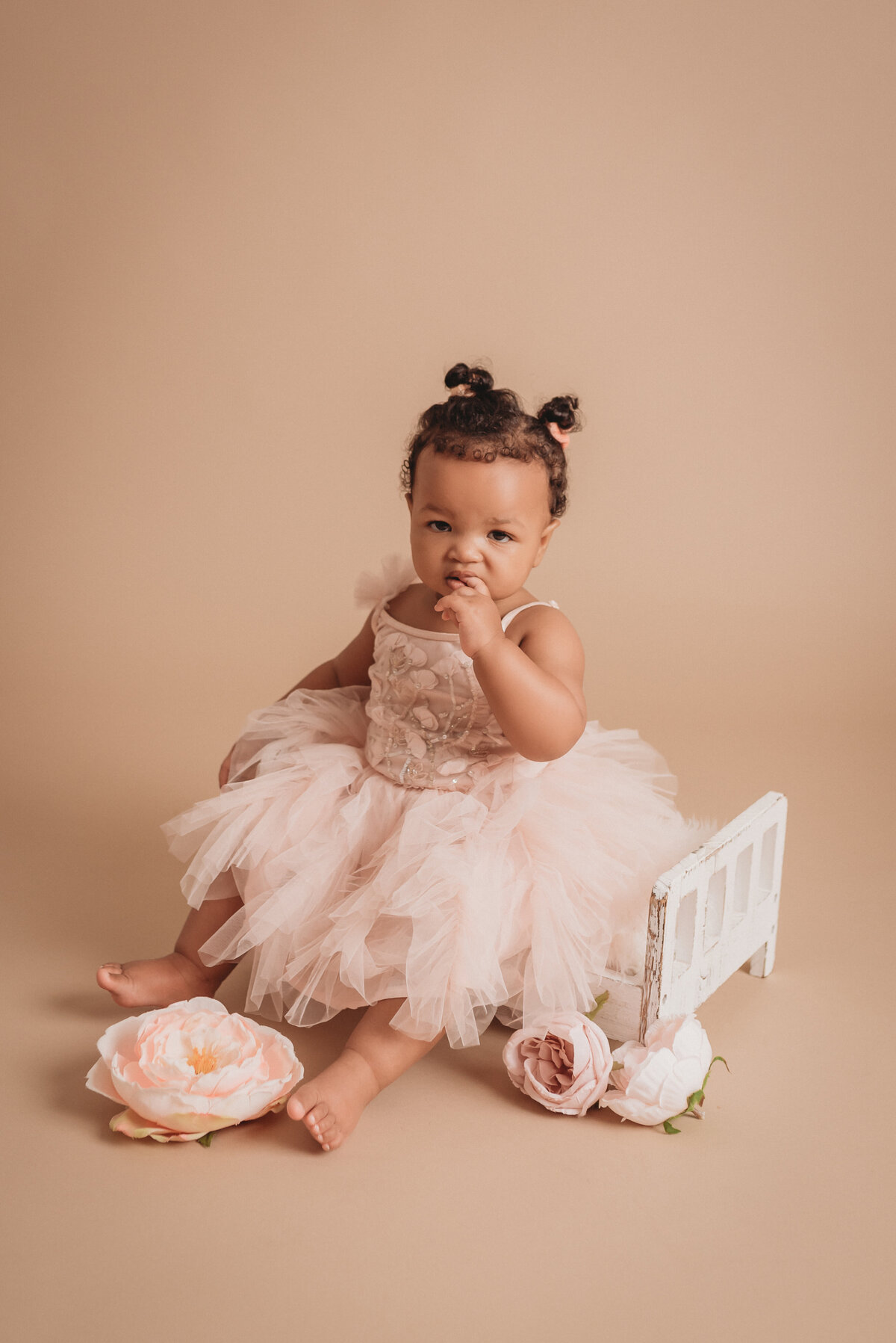 One year old baby girl in pink tulle dress sitting on miniature white bed with pink flowers around her on tan backdrop