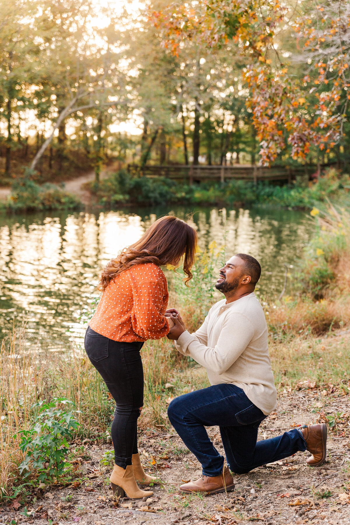 A man is on his knee proposing with a beautiful pond and trees in the background.