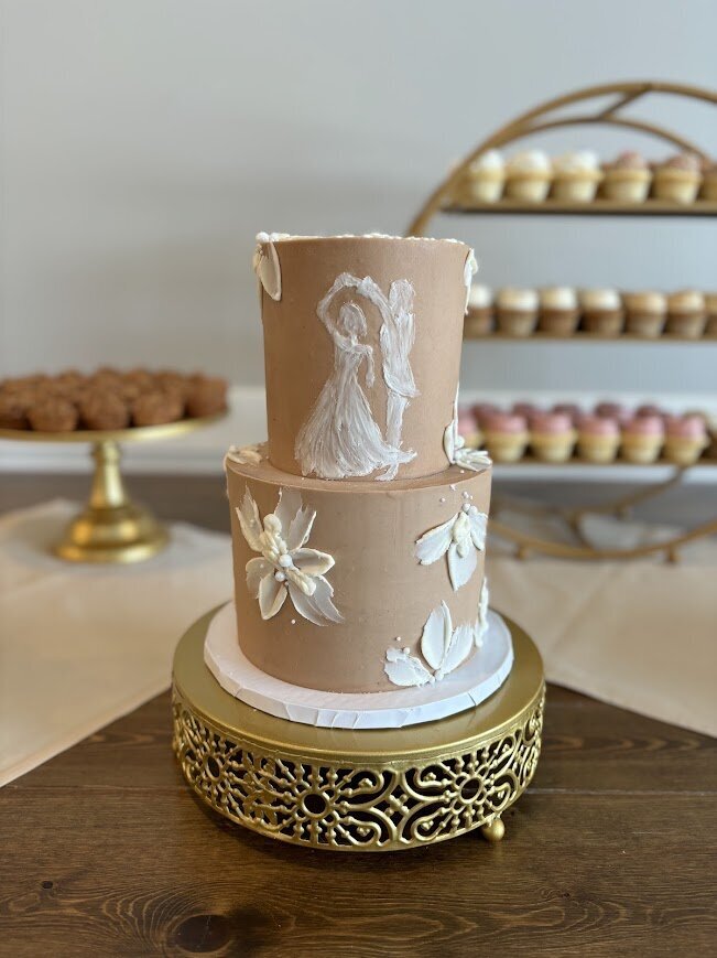 A two tier cake sits in the foreground of a table on a gold stand. The cake has chocolate frosting with white buttercream painted flowers, sugar pearls, and a white painted silhouette of a couple dancing together. In the background are slightly blurred desserts on other gold stands - mini cupcakes and peach pies.