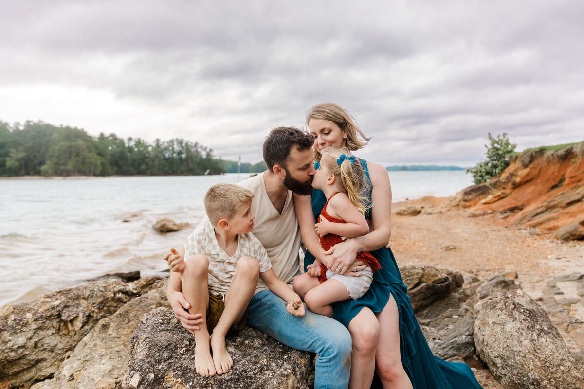 Family of 4 sits on rock at lake and Dad kisses young daughter