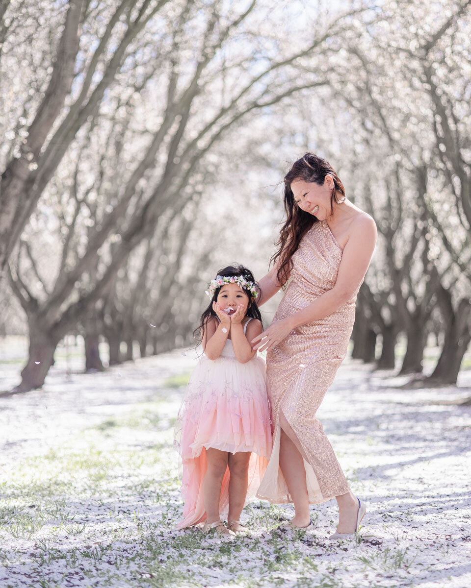 Four-year-old daughter blowing almond blossom petals from her palm while mom watches lovingly during spring photoshoot in blooming orchard