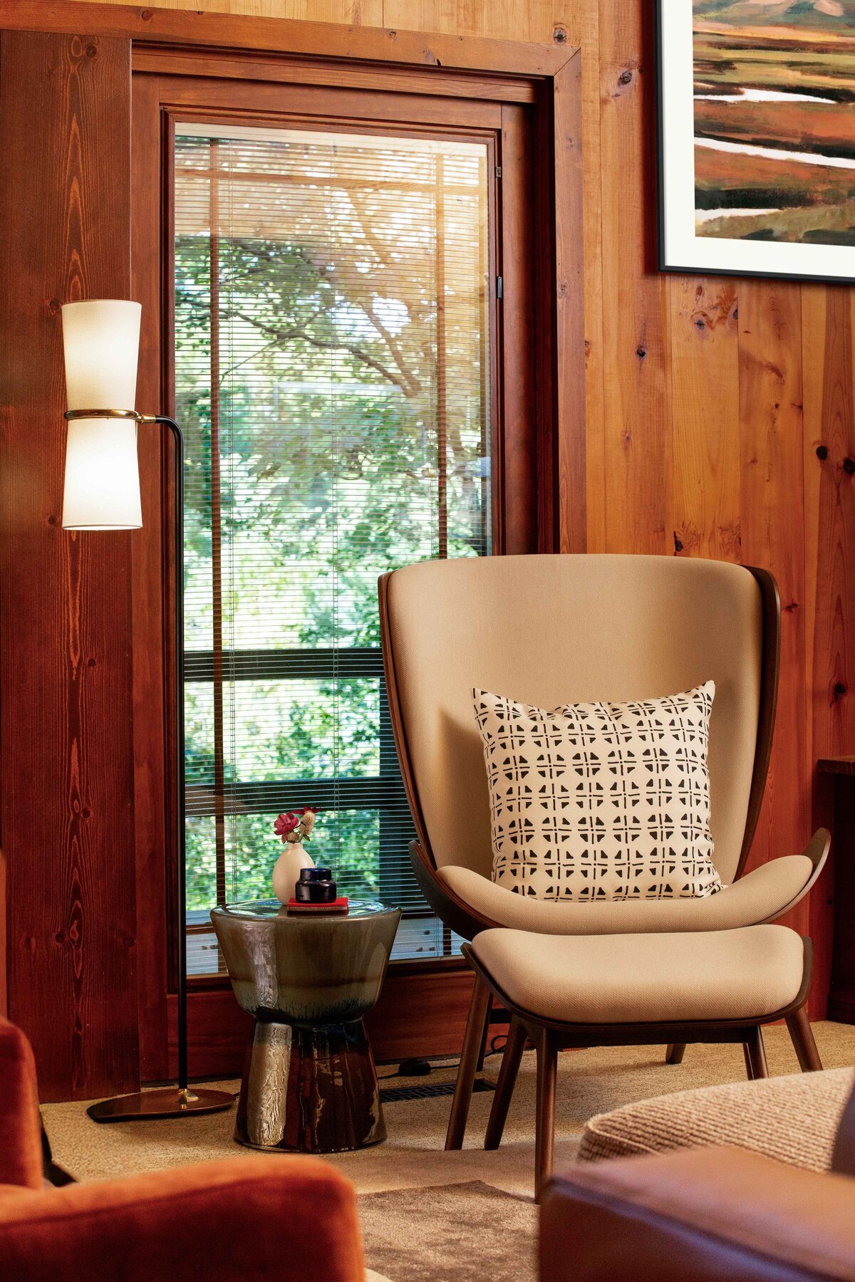A cozy reading nook in a wood cabin. A tan armchair with a patterned pillow sits next to a small, textured side table holding a vase with flowers and a book. A modern floor lamp with two cylindrical shades stands behind the table. Sunlight filters through a window with blinds, offering a view of tree