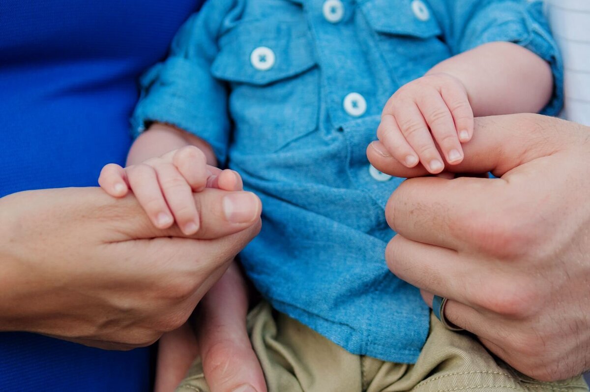 The image shows a close-up of three hands. A child's small hands are holding the thumbs of two adult hands, one on each side. The child is wearing a blue shirt with white buttons and beige pants. One of the adults is wearing a blue garment, and the other adult has a silver ring on their finger. The focus is on the hands and the child's torso.