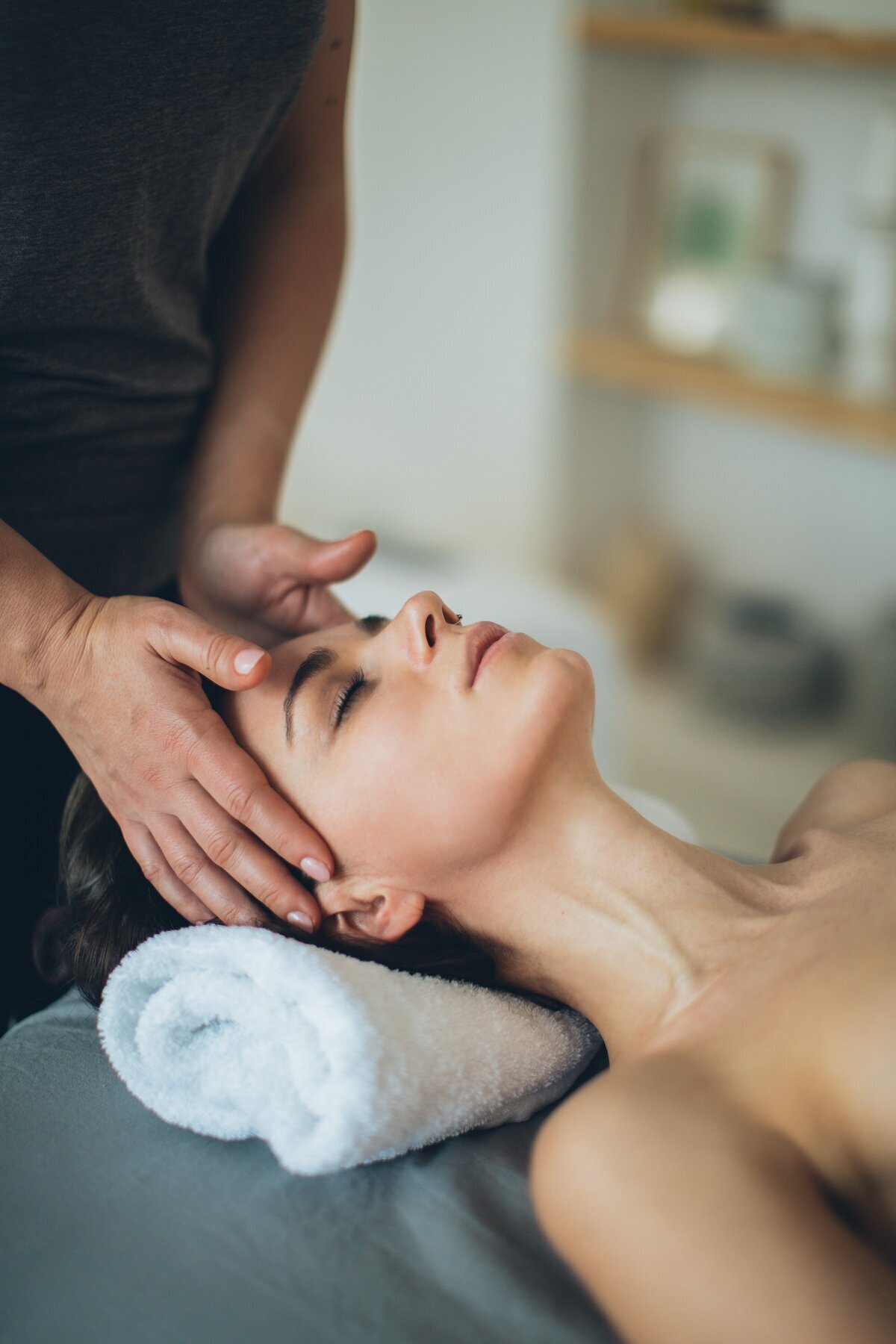 Visage d'une femme allongée, yeux fermés, tête reposant sur un drap en train de se faire masser le crâne.