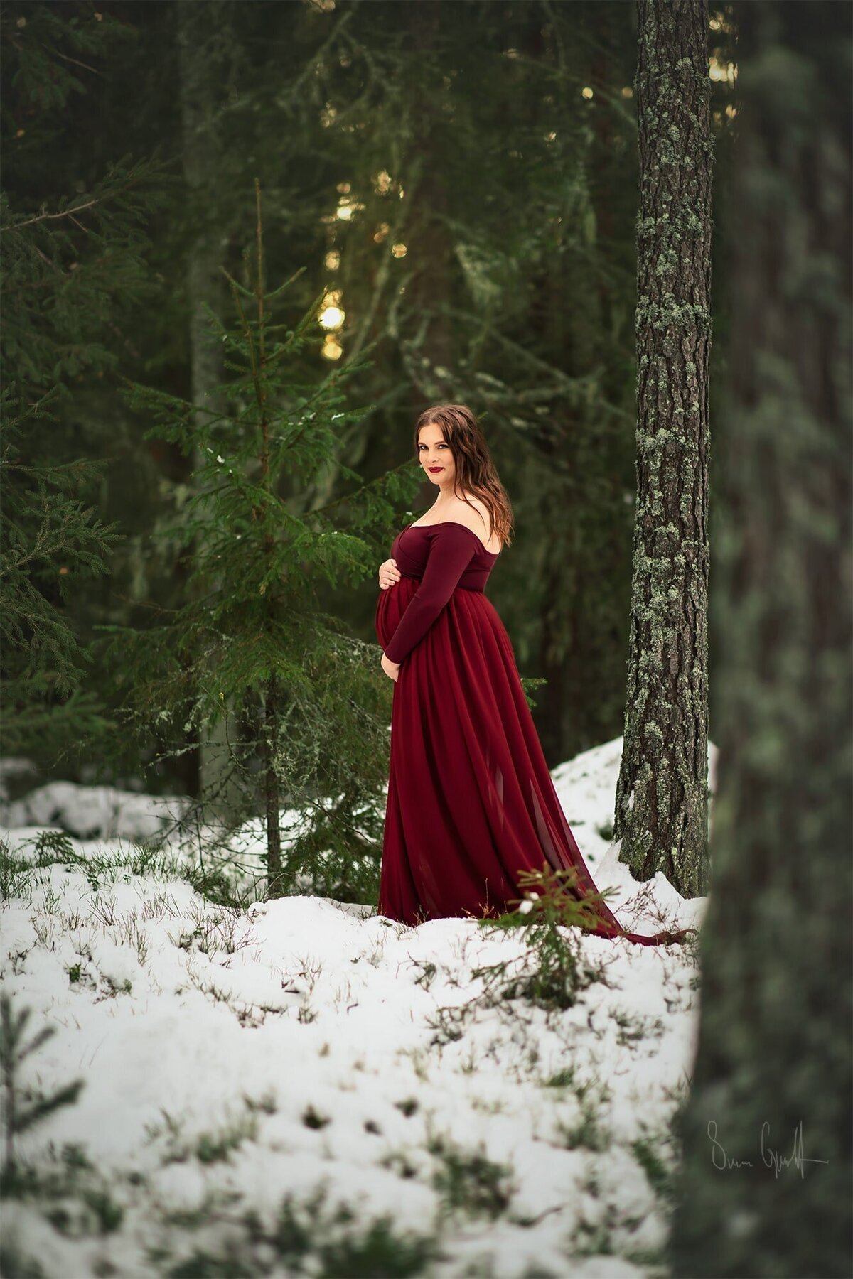 konstnärlig fine art fotografering av gravid utomhus. Gravid kvinna i röd klänning står i skogen och håller händerna på magen.