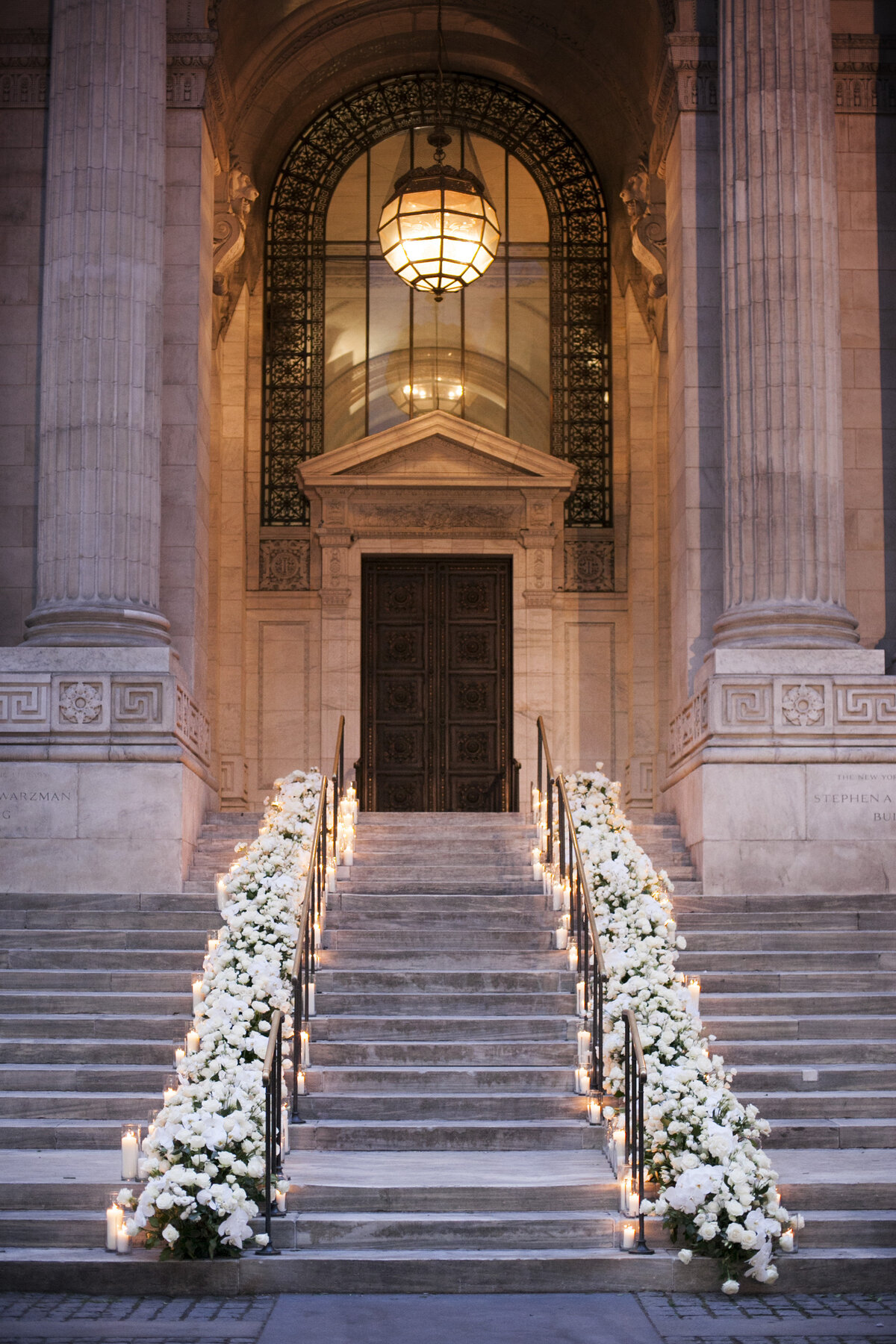 A grand stone staircase leading up to an ornate building entrance decorated with a floral arrangement of white flowers and candles. The entrance features large columns, an arch with intricate metalwork, and a large hanging lantern.